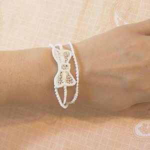 Puede incluir: Pulsera de encaje blanco con un diseño de lazo. La pulsera está hecha de encaje blanco delicado y presenta un diseño en forma de lazo en el centro. La pulsera se muestra en la muñeca de una persona.