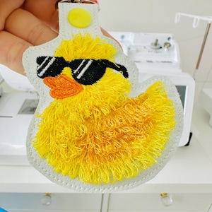 May include: A yellow duck keychain with black sunglasses and an orange beak. The duck is embroidered with yellow thread and has a white border. The keychain has a yellow button and a silver ring.