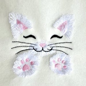 Op de afbeelding: Wit geborduurd kattengezicht met roze oren en poten. De kat heeft een glimlachende uitdrukking met gesloten ogen.