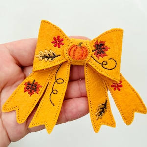 May include: Mustard yellow felt hair bow with embroidered fall-themed accents. The bow features a central pumpkin design and decorative leaves and floral patterns. A festive accessory for autumn.