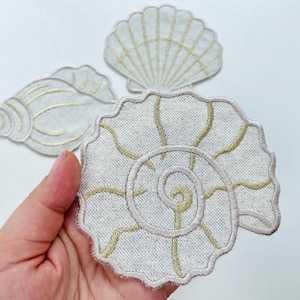 以下が含まれることがあります： さまざまな形の刺繍された貝殻パッチが3つ。パッチは、金色のステッチが施された薄いベージュ色の生地で作られています。最大のパッチはらせん状の貝殻で、その他は扇形と巻貝形です。