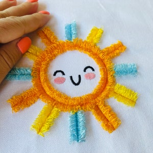 May include: A yellow embroidered sun with a smiling face and blue and yellow rays. The sun is on a white background.