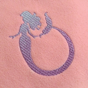 May include: A pink fleece blanket with a purple embroidered mermaid design. The mermaid is curled in a circle with her tail forming the circle.