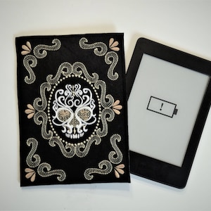 May include: A black e-reader with a low battery warning icon on the screen. The e-reader is lying on a white surface next to a black fabric cover with an embroidered sugar skull design.