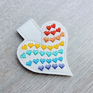 May include: White heart-shaped bookmark with a rainbow of embroidered hearts. The bookmark has a small white tab at the top for insertion into a book. The embroidered hearts are in rows of red, orange, yellow, teal, blue, and purple.