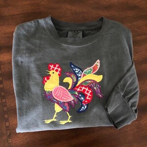 May include: A gray long-sleeved t-shirt with a colorful rooster applique. The rooster is made of fabric scraps in shades of yellow, pink, purple, blue, and green.