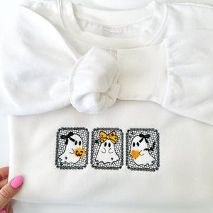 May include: A white sweatshirt with three embroidered ghost designs. Each ghost has a black bow and is holding a pumpkin or heart. The ghosts are framed in black lace. The sleeves are folded over the front of the sweatshirt.