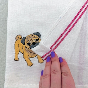 May include: White hand towel with a detailed embroidered pug dog. The towel has red stripes and is folded to reveal the pug design. The pug is tan, black, and brown.