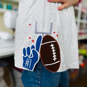 May include: A gray t-shirt with an embroidered football, goal post, and a blue number one hand. The hand is pointing up and the football is brown with white stitching.