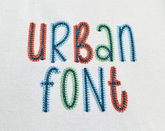 May include: Embroidered text on white fabric reads "URBAN FONT". The letters are in a playful, stitched style with a combination of blue, green, and orange thread. The background is a plain white fabric.