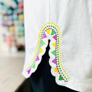 May include: White t-shirt with a colorful embroidered design on the side seam. The design features purple, green, and yellow triangles and dots.