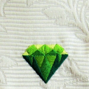 Puede incluir: Un parche bordado en forma de diamante verde sobre un fondo de tela blanco.