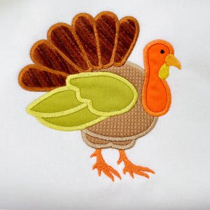 May include: Embroidered turkey applique in shades of brown, orange, and green on a white background. The turkey has a detailed design with a textured body, a fan of brown feathers, and orange legs and beak. A festive Thanksgiving decoration.