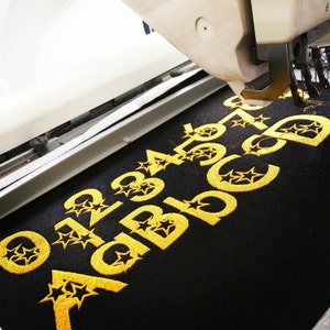 以下が含まれることがあります： 黒い布地に、金色の刺繍で数字の01234567と「ABCDE」という単語が刺繍されています。各文字と数字の周りを星が囲んでいます。