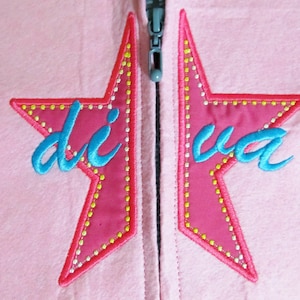 May include: Pink zippered jacket with two embroidered stars, each with a yellow outline and a blue inscription. The left star says "diva" and the right star says "di", creating the word "diva" when the stars are together.