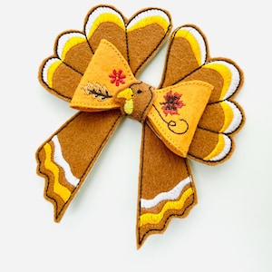 May include: A felt turkey bow in shades of brown, yellow, and white. The bow has a turkey head and tail feathers, with embroidered details of a flower and leaf. Ideal for Thanksgiving or autumn-themed accessories.