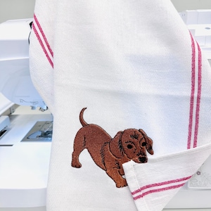 May include: White kitchen towel with a brown dachshund embroidery. The towel has two vertical red stripes and is folded over. The dog is embroidered in a detailed, realistic style.