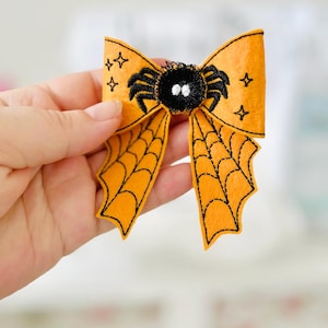 May include: Orange Halloween bow with a black spider and web design. The bow features a felt spider with white eyes and embroidered details. The bow's ends have a spider web design and star accents.