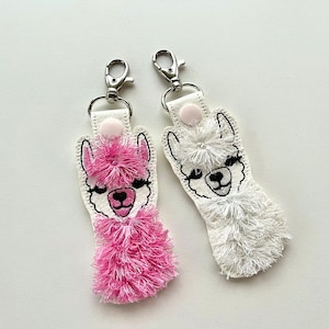 May include: Two llama keychains, one with pink fur and one with white fur. Both have black eyes and a black nose.