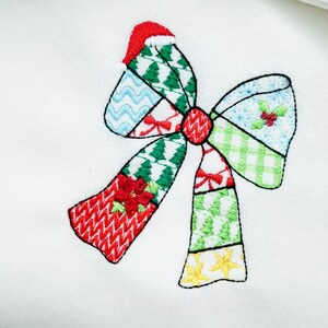 K&ouml;nnte beinhalten: Gestickte Weihnachtsschleife mit Patchwork-Design. Die Schleife hat rote, gr&uuml;ne, blaue und wei&szlig;e Abschnitte mit festlichen Mustern wie Weihnachtsb&auml;umen, Stechpalmen und Sternen. Die Schleife ist vor einem wei&szlig;en Hintergrund platziert.