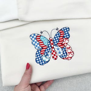 May include: A folded, cream-coloured fabric item with an embroidered butterfly design. The butterfly's wings are a patchwork of red, white, and blue patterns, including stars, checks, and stripes. The item is held by a hand with red nail polish.