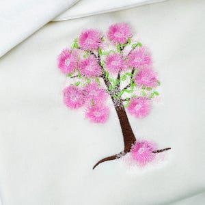 Puede incluir: Diseño bordado de un árbol en flor con flores rosas, hojas verdes y un tronco marrón. El árbol está sobre un fondo blanco. Las flores tienen una textura esponjosa.