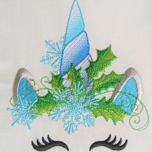 May include: Embroidered design of a unicorn's face with a blue horn, green holly leaves, and blue snowflakes. The unicorn has silver ears and black eyelashes.