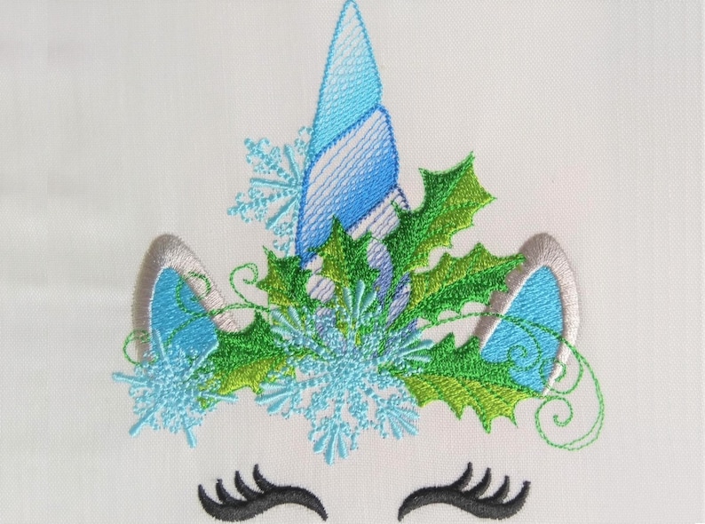 May include: Embroidered design of a unicorn's face with a blue horn, green holly leaves, and blue snowflakes. The unicorn has silver ears and black eyelashes.