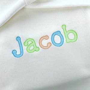 May include: White baby blanket with the name "Jacob" embroidered in colorful thread. The letters are outlined in blue, green, and orange, creating a playful and personalized design. The blanket is folded.