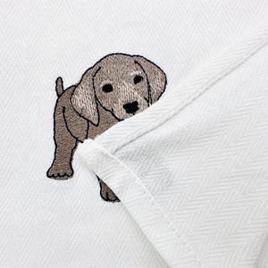 May include: Embroidered image of a light brown puppy on a white fabric with a herringbone pattern. The puppy is facing forward with its head turned slightly to the side. The fabric is folded over, creating a layered effect.