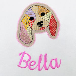 May include: Embroidered dog face applique with a patchwork design in tan, red, yellow, and purple. The applique is outlined with a pink lace trim. The name "Bella" is embroidered in pink script below the dog.