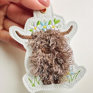May include: A white felt ornament featuring an embroidered brown Highland cow wearing a flower crown. The cow is standing on a green grass patch.