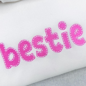 May include: White fabric with the word "bestie" embroidered in bright pink. The letters are outlined with white stitching, creating a playful and textured appearance. The design is simple and eye-catching.