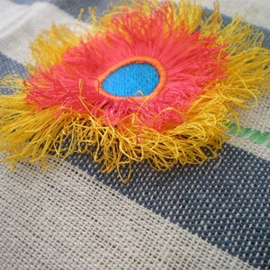 May include: A yellow, red, and blue embroidered flower with a fringed edge. The flower is on a striped fabric background.