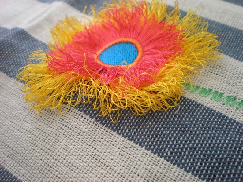 May include: A yellow, red, and blue embroidered flower with a fringed edge. The flower is on a striped fabric background.