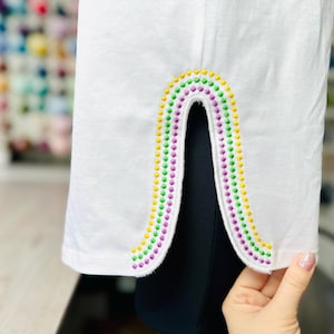 May include: White t-shirt with a colorful rainbow design sewn on the back. The rainbow is made of green, yellow, and purple beads.