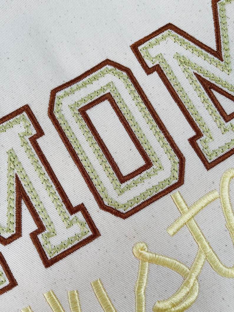 May include: A beige fabric with brown and green embroidered lettering that reads "MON" and a yellow embroidered design that reads "CUSTOM".