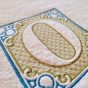 May include: A white towel with a blue and green embroidered monogram of the letter "O" in a decorative frame.