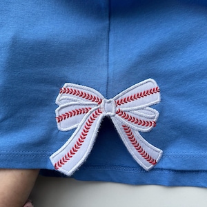 以下が含まれることがあります： 薄い青色のTシャツに白いボウのアップリケが付いている。ボウは野球のステッチのような赤いステッチが施されている。