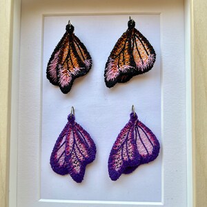 May include: Four butterfly-shaped earrings displayed in a white frame. Two earrings are black, orange, and pink, and the other two are purple and pink.