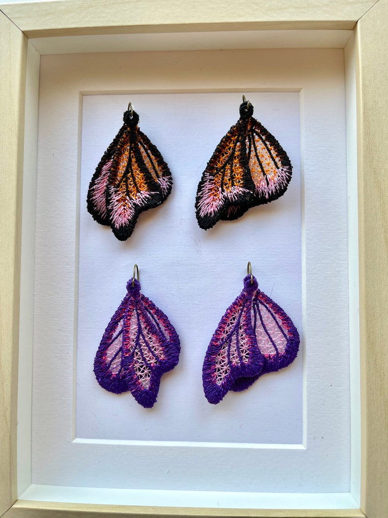 May include: Four butterfly-shaped earrings displayed in a white frame. Two earrings are black, orange, and pink, and the other two are purple and pink.