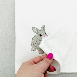 Puede incluir: Toalla de mano blanca con un bordado de conejo gris con detalles en rosa. La toalla se levanta para revelar el diseño. El conejo está representado de pie.