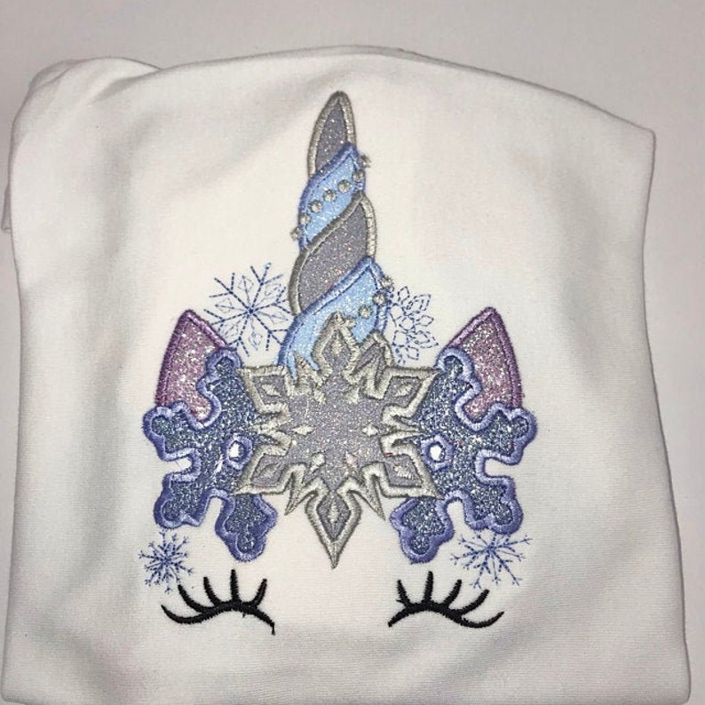 Op de afbeelding: Witte geborduurde trui met een sneeuwvlok-eenhoorn ontwerp. De eenhoorn heeft een blauwe en zilveren spiraalvormige hoorn, paarse oren en sneeuwvlok details.