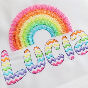 May include: Embroidered rainbow with a zig-zag pattern in multiple colors, spelling out the name "Lucia"