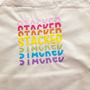 May include: A white canvas tote bag with the word "STACKED" embroidered in rainbow colors. The word is repeated multiple times, with each repetition getting smaller and moving slightly to the right.