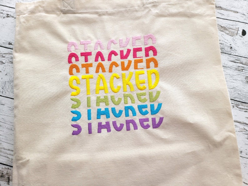 May include: A white canvas tote bag with the word "STACKED" embroidered in rainbow colors. The word is repeated multiple times, with each repetition getting smaller and moving slightly to the right.