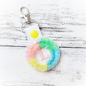 May include: A white keychain with a yellow button and a colorful circle with the letter L embroidered in pink. The circle is surrounded by a pink, yellow, and blue fringe.