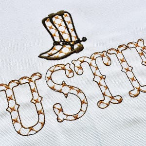 Può includere: Nome ricamato "JUSTIN" in un motivo a punto croce marrone con un disegno di stivale da cowboy marrone sopra il nome.