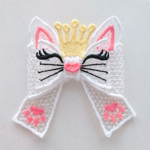 Puede incluir: Lazo de encaje blanco con una cara de gato rosa, pestañas negras, una nariz rosa y una corona amarilla. El lazo tiene huellas de patas rosas en la parte inferior.
