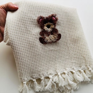 Könnte beinhalten: Ein weißes Waffelgewebe-Handtuch mit einem gestickten Teddybär-Motiv in Braun und Beige. Das Handtuch hat einen weißen Fransenrand.
